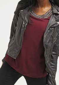 Giacca in pelle nera da motociclista con dettagli in argento, indossata sopra un top bordeaux con un colletto di perline. Texture liscia, design aderente.