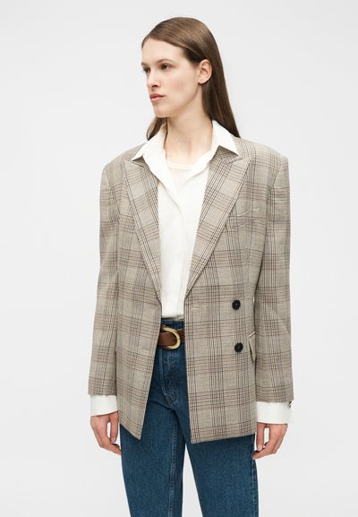 Frau trägt einen beigen, karierten, doppelt geknöpften Blazer über einem weißen Hemd und blauen Jeans, steht vor einem schlichten weißen Hintergrund.