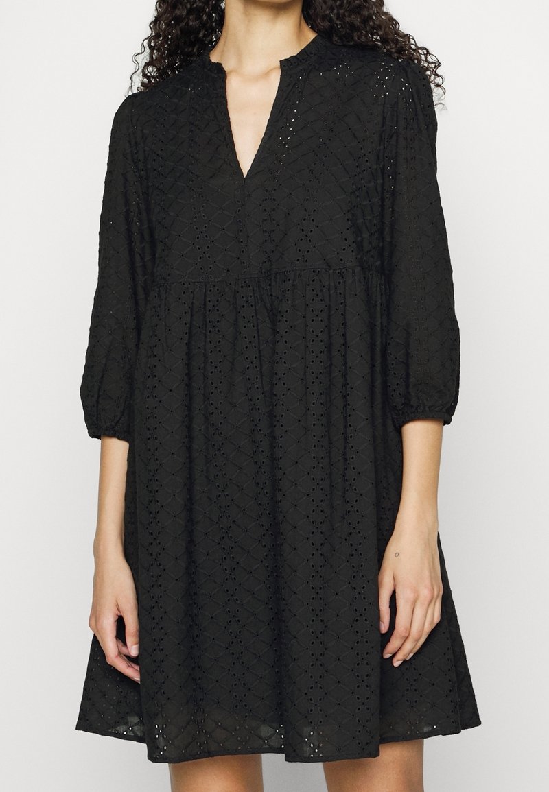 Femme portant une robe noire longueur genou avec des manches ballon trois-quarts et des motifs losanges brodés ajourés.