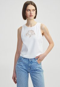 Witte katoenen mouwloze top met een bloemenkant applicatie in het midden, gecombineerd met lichtblauwe spijkerbroek met witte stippen.
