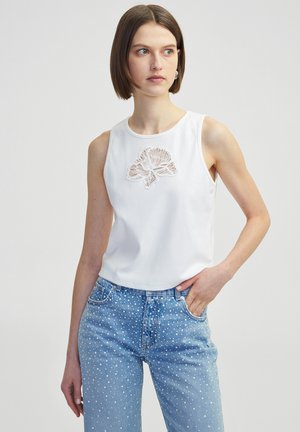 Witte katoenen mouwloze top met een bloemenkant applicatie in het midden, gecombineerd met lichtblauwe spijkerbroek met witte stippen.