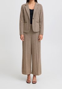 Giacca da suit a motivo herringbone marrone e pantaloni a gamba larga, realizzati in materiale testurizzato. Abbinata a un top nero e sandali neri con cinturino.