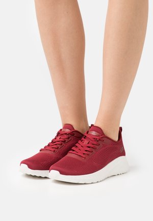 Personne portant des baskets en tricot rouges avec des semelles blanches, debout sur une surface blanche.