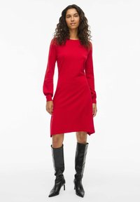 Rotes langärmliges Kleid mit geradem Schnitt, aus weichem Stoff. Getragen mit hohen schwarzen Stiefeln. Einfaches Design, keine sichtbaren Muster oder Akzente.