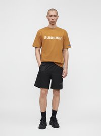 Un homme est debout, face à l’avant, portant un t-shirt marron "SUNBURN", un short noir, des chaussettes noires et des baskets noires sur un fond uni.