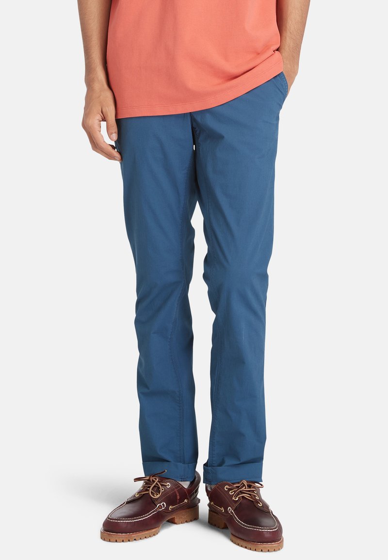 Lichtblauwe broek van een gladde, lichte stof. Heeft een rechtlijnig ontwerp en wordt gecombineerd met donkerenbruine schoenen.