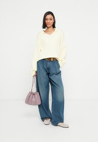 Μοντέλο που φοράει ένα υπερμεγέθες πουλόβερ φωτεινού κίτρινου χρώματος, μπλε τζιν παντελόνι με φαρδιά εφαρμογή, γκρι παπούτσια τύπου slip-on και κρατά μια ανοιχτό μωβ τσάντα με κορδόνι.