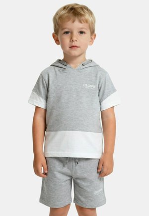 Jeune garçon aux cheveux blonds portant un t-shirt à capuche à manches courtes gris et blanc, accompagné d'un short assorti, se tenant devant un fond uni.