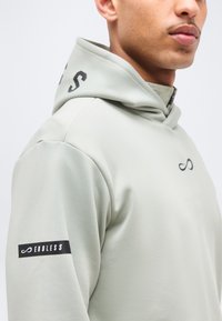 Homme portant un sweat à capuche gris clair avec le texte noir "ENDLESS" et des symboles d'infini sur la poitrine, la manche et la capuche.