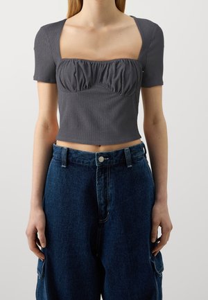 Femme portant un haut court gris à manches courtes et col carré avec un détail froncé, associé à un pantalon baggy en denim bleu foncé taille haute.