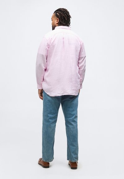 Άνδρας με πλεξούδες, που φοράει ανοιχτό ροζ μακρυμάνικο πουκάμισο, μπλε τζιν και καφέ παπούτσια, στέκεται με την πλάτη προς το μέρος μας σε λευκό φόντο.