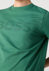 Vihreä lyhythihainen t-paita, jossa on hienovaraisesti kohokuvioitu "BOSS" -logo rinnan kohdalla, käytössä henkilö, joka on kuvattu chinin alapuolelta.