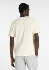 T-shirt en coton couleur crème à manches courtes, avec un col rond et une coupe décontractée. Porté avec un pantalon de survêtement gris.