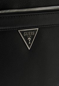 Section d'un sac en cuir noir texturé avec une fermeture éclair argentée et un logo triangulaire en métal de Guess USA avec un point d'interrogation au centre.