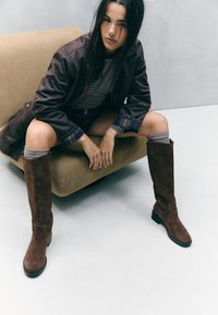 Bruna mockaskinnknähöga stövlar med rund tå och platt klack. Modellen bär en randig tröja, shorts och grå knästrumpor, sittande på en beige soffa.