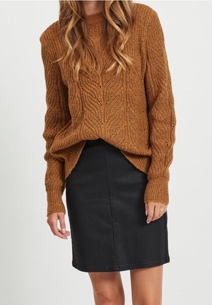Pull en maille torsadée marron associé à une jupe noire longueur genou sur une personne debout aux cheveux longs et ondulés.