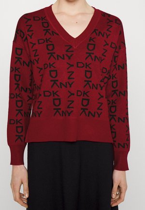 Žena v červeném svetru s dlouhými rukávy a výstřihem do V, s černým vzorem loga DKNY, a v černé sukni na jednobarevném pozadí.