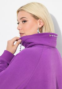 Lila Kapuzenpullover mit hohem Kragen und Kordelzug, ausgestattet mit gerippten Bündchen. Glatte Textur mit dezenten Nähtestichen. Keine sichtbaren Logos.
