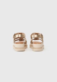 Rückansicht eines goldenen Metallic-Sandalen mit mehreren Riemen, verstellbarem Schnallenverschluss und einer hellbraunen Plateausohle mit strukturierten Details.