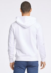 Sudadera blanca con capucha, con puños y dobladillo acanalados. Textura suave y corte relajado, se muestra combinada con pantalones azules texturizados.