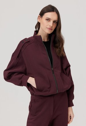 Kobieta w ciemnoczerwonej kurtce na zamek z bufkami na rękawach i dopasowanych spodniach, ręce częściowo w kieszeniach, na jasnoszarym tle.