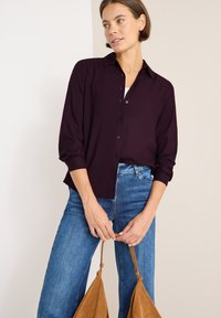 Mørk lilla skjorte med krave, lange ærmer og afslappet pasform, parret med blå jeans og en brun taske. Glat stofstruktur.
