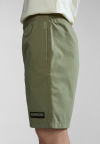 Person wearing light green casual shorts with an elastic waistband and a black rectangular logo patch near the hem on the side.