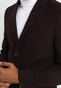 Blazer burdeos oscuro con un solo botón negro, hecho de una tela suave. Presenta solapas con muesca y un corte entallado, usado sobre un cuello de tortuga blanco.