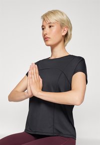 Črna, kratkosleeve majica z teksturo za šport z okroglo ovratnikom; model sedi, roke v položaju molitve, oblečen v bordo legice.