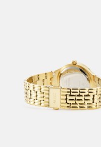 Goldener Metalluhren mit einem verlinkten Armbanddesign und einem runden Gehäuse. Besitzt ein strukturiertes Band und eine silberne Rückseite mit Markenprägung. Kreisförmige Krone.