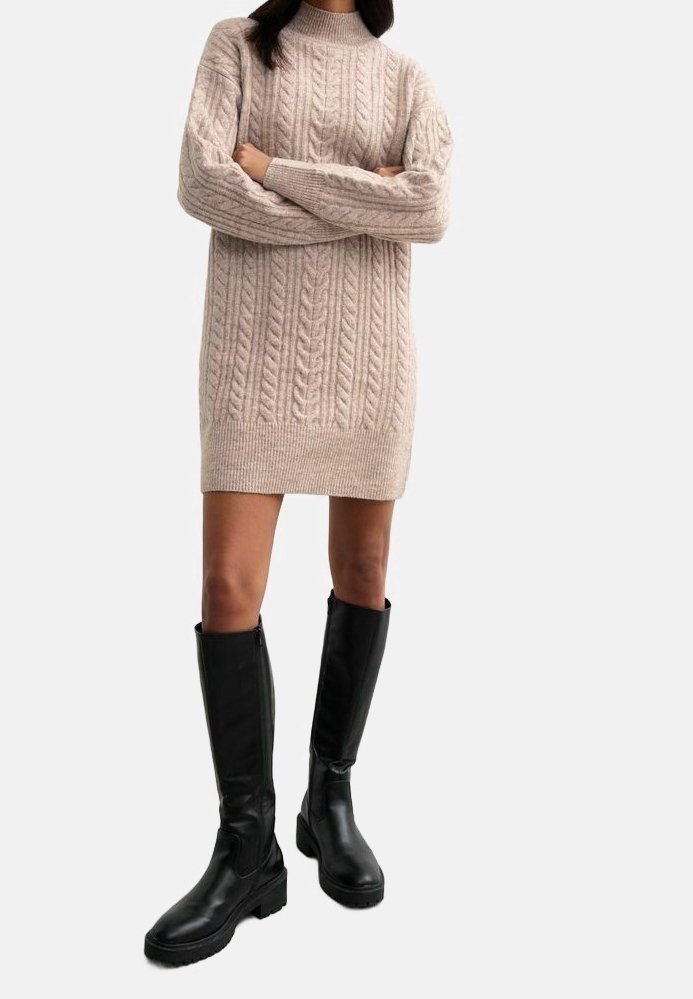 Žena v béžových šatech s pleteným vzorem a černými koženými kozačkami po kolena, stojící se zkříženýma rukama.