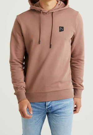 Mand iført en støvet pink hættetrøje med snører og en lille sort logo på brystet, kombineret med lyseblå jeans.