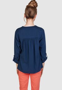 Navyblaue Bluse mit einem plissierten Rücken, drei Viertelärmel hochgekrempelt, kombiniert mit korallfarbenen Hosen. Glatte Stofftextur.