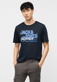 Jaunas vyras su juodais garbanotais plaukais, dėvintis tamsiai mėlyną „Jack & Jones Superior New Aesthetics“ marškinėlį ir pilkas kargo kelnes, rankomis kišenėse.