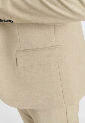 Veste de costume beige en tissu texturé, avec une poche arrière unique et des lignes épurées et ajustées. Les boutons sur le poignet sont visibles.