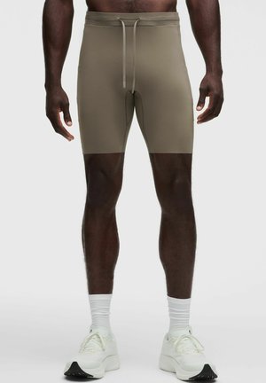 Mann trägt beige enganliegende Shorts mit Kordelzug, weiße Kniestrümpfe und weiße Sportschuhe, steht vor hellgrauem Hintergrund.