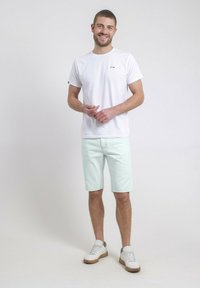 T-shirt blanc à col rond avec un petit logo, shorts en denim vert menthe et baskets blanches avec semelles beiges. Le modèle se tient devant un fond neutre.