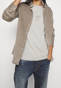 Ljust brunt kordflorskjorta som är lager på lager över en ljusgrå t-shirt med en skissad design. I kombination med blå denimjeans ger detta en avslappnad look.