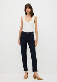 Camiseta de tirantes blanca acanalada combinada con jeans de mezclilla azul oscuro que cuentan con bolsillos laterales y cierre de botón, rematada con zapatos de tacón marrón claro.