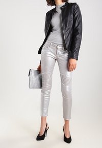 Veste en cuir noir sur un haut gris, jeans skinny argentés brillants avec détails texturés, et escarpins noirs. Tenant une pochette métallique.