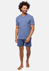 Kortärmad, blå och vitrandig t-shirt med V-hals och ficka, kombinerad med ensfärgade blå shorts. Barfota, stående mot en vit bakgrund.