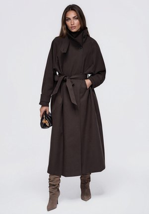 Une femme porte un long manteau marron foncé, ceinturé et à col montant, des bottes en daim montant jusqu’aux genoux et un petit sac à main noir.
