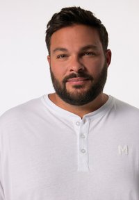Maglietta henley bianca a maniche corte con tre bottoni e un piccolo logo sul petto. Tessuto liscio con una vestibilità rilassata e informale.