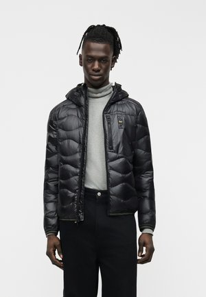 Noor tumedama nahaga noormees, kellel on dredi-soeng, kannab musta puffer-jakki helehallile kõrge kaelusega kampsunile ja mustadele pükstele.