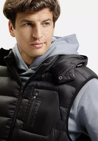 Mężczyzna w jasnoniebieskiej bluzie z kapturem, noszący bezrękawnik w kolorze czarnym z zamkiem błyskawicznym na klatce piersiowej, patrzy nieco w lewo.