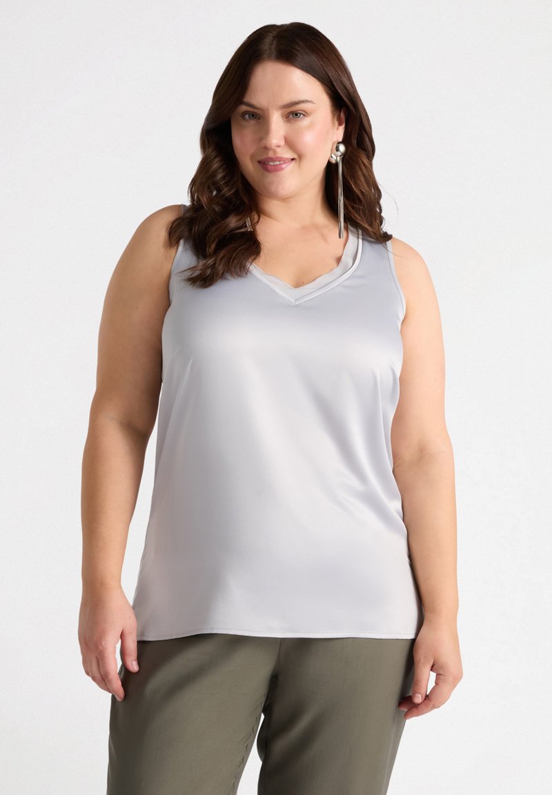 Vaaleanharmaa hihaton toppi, jossa on V-pääntie ja sileä, kiiltävä kangas. Yhdistetty vihreisiin, löysiin housuihin.