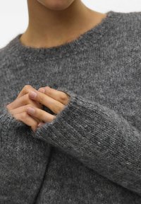 Pull en tricot gris avec une finition texturée et des poignets larges, bien ajusté au niveau du cou, porté avec les mains jointes.