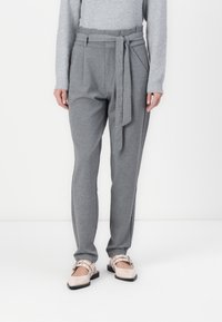Pantalons gris avec une taille à nouer, présentant une coupe décontractée et des poches latérales. Accompagnés de chaussures claires et brillantes.