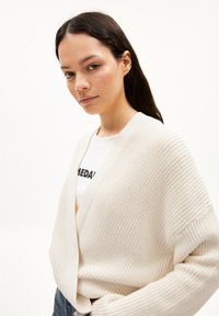 Beige gebreide cardigan met een V-hals ontwerp, dikke ribbeltekstuur en open voorkant. Draag het over een wit T-shirt met zwarte print.