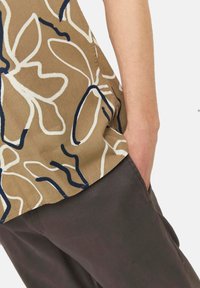 Chemise beige avec un motif florale abstrait blanc et bleu marine, présentant une coupe décontractée, des manches courtes et un ourlet arrondi. Associée à un pantalon foncé.